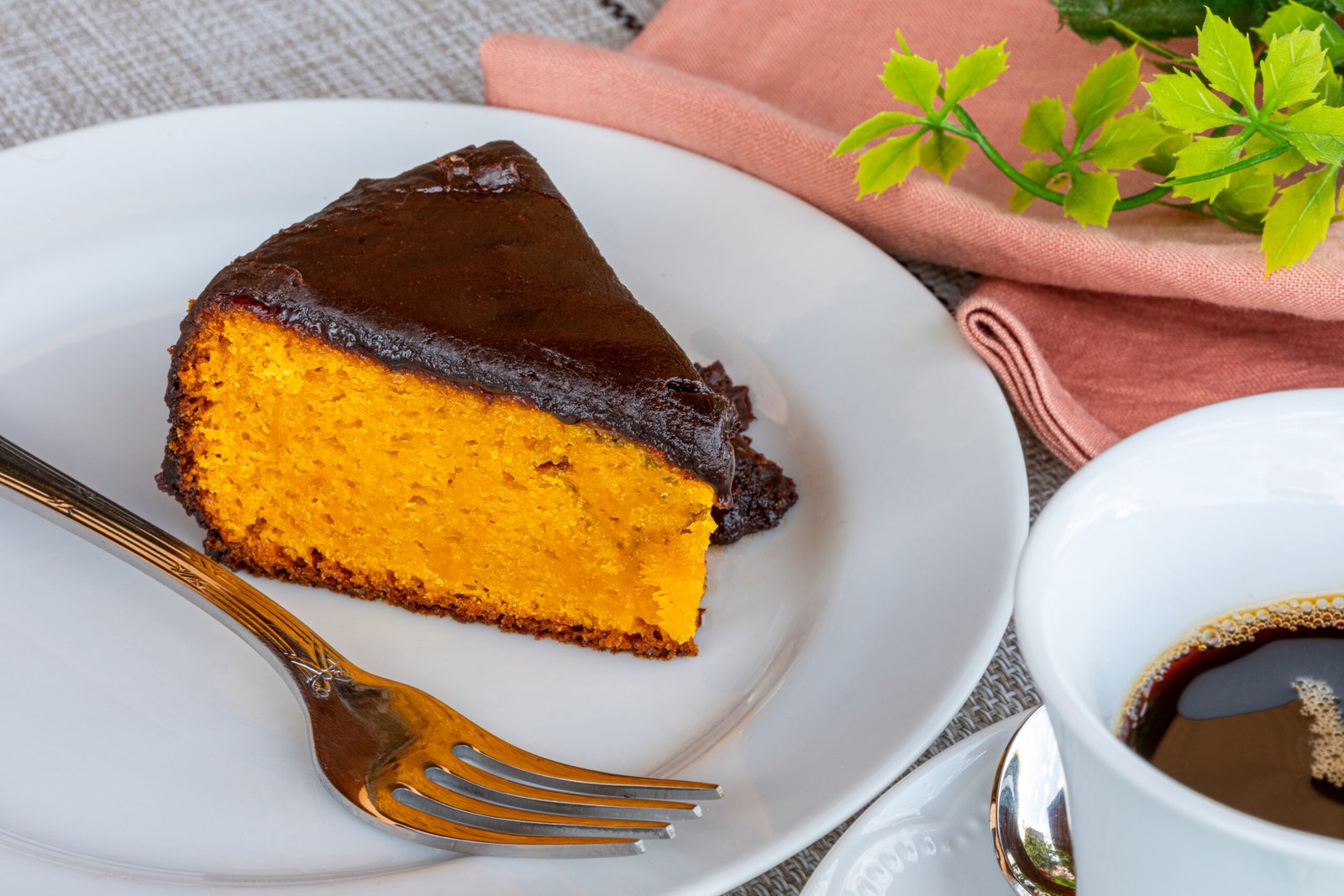 Fatia de bolo de cenoura com cobertura de chocolate servido em um prato branco, acompanhado de uma xícara de café