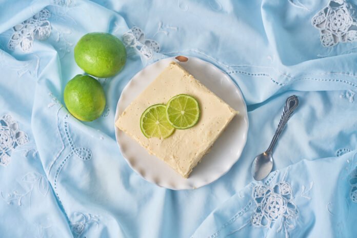 Fatia de bolo gelado de limão com leite de coco decorada com rodelas de limão sobre prato branco.