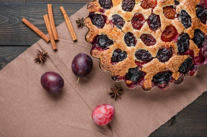 Bolo de ameixa com canela em uma travessa decorativa, acompanhado de paus de canela e ameixas frescas sobre um pano marrom.