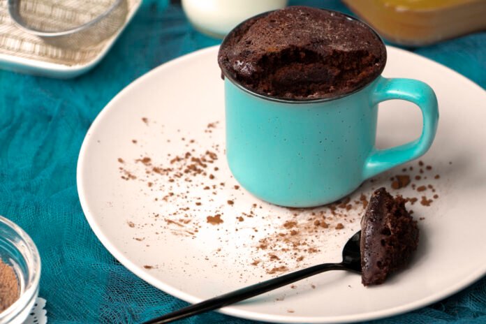 Bolo de caneca simples de chocolate servido em uma caneca azul com uma colher ao lado, sobre um prato branco, em uma mesa azul.