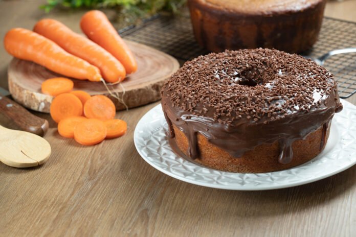 Bolo de cenoura fofinho com cobertura de brigadeiro, decorado com granulado de chocolate em um prato branco, acompanhado de cenouras frescas ao fundo.