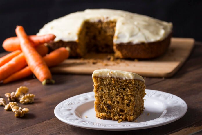 Fatia de bolo de cenoura vegano com cobertura cremosa, servida em um prato branco, ao lado de cenouras e nozes.