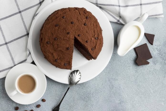 Bolo de chocolate vegano e sem glúten em prato branco, com uma fatia retirada ao lado de xícara de café e pedaços de chocolate.