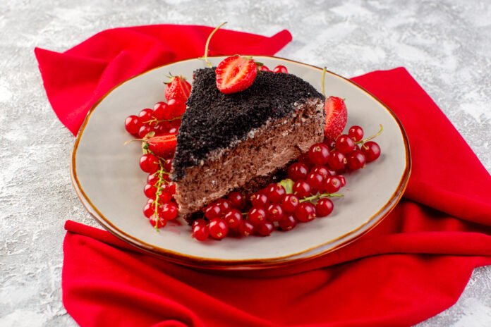 Fatia de bolo de chocolate decorado com frutas vermelhas frescas, morangos e groselhas em um prato com fundo vermelho.