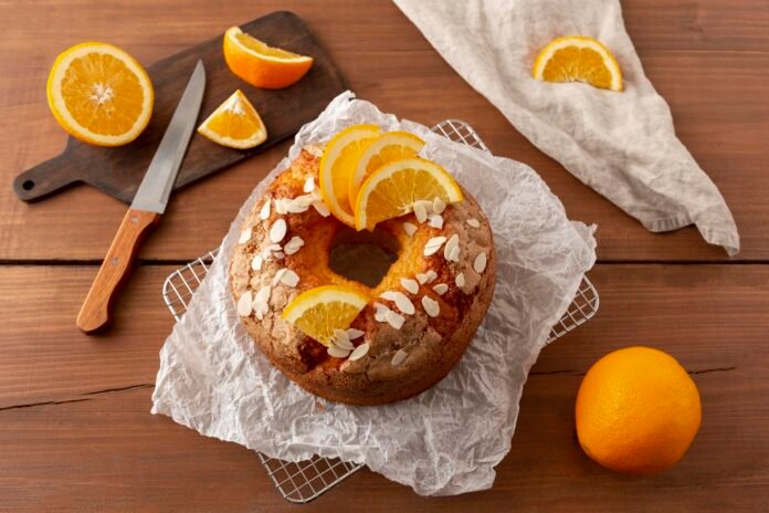 Bolo de laranja integral decorado com amêndoas e fatias de laranja sobre papel manteiga.