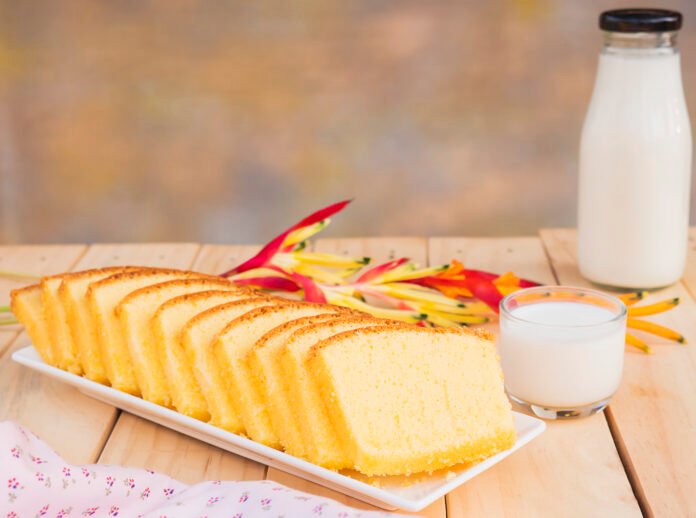 Fatias de bolo de leite condensado cremoso e fácil em um prato branco, com flores coloridas ao fundo e um copo de leite ao lado.