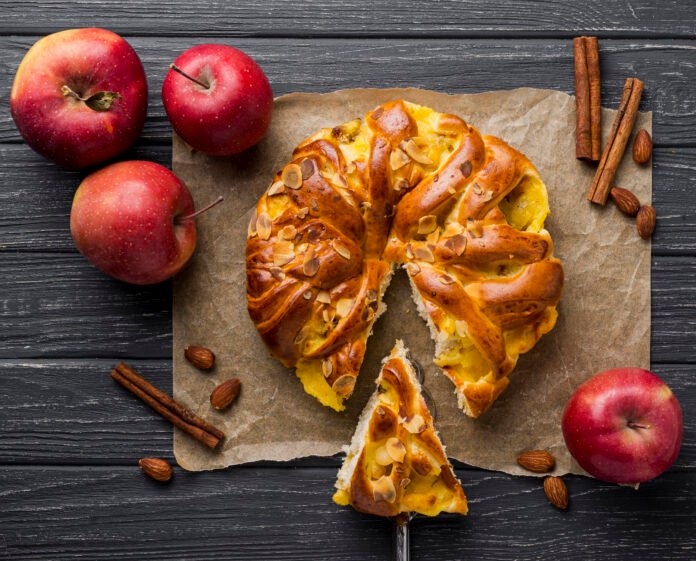 Bolo de maçã e canela decorado com amêndoas em uma mesa com maçãs e paus de canela.