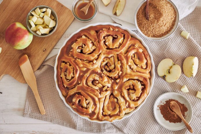 Rolinhos de maçã e canela com cobertura caramelizada em prato branco, com ingredientes como maçãs picadas e açúcar mascavo ao redor.