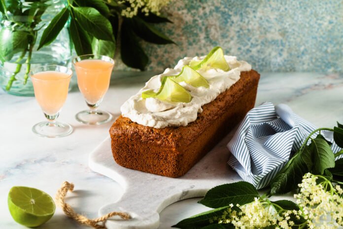 Bolo de limão com cobertura cremosa de chantilly e fatias de limão como decoração, acompanhado por duas taças de suco em um ambiente decorado.