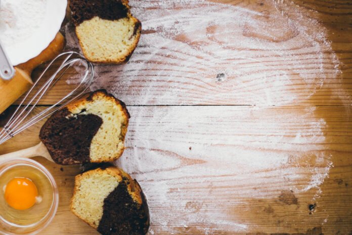 Fatias de bolo mesclado com utensílios e ingredientes sobre uma mesa de madeira, destacando simplicidade e rusticidade.