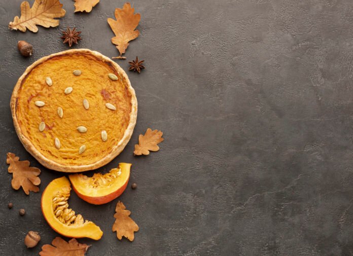 Bolo de abóbora vegano decorado com sementes de abóbora, ao lado de fatias frescas de abóbora e folhas secas em uma superfície rústica escura.