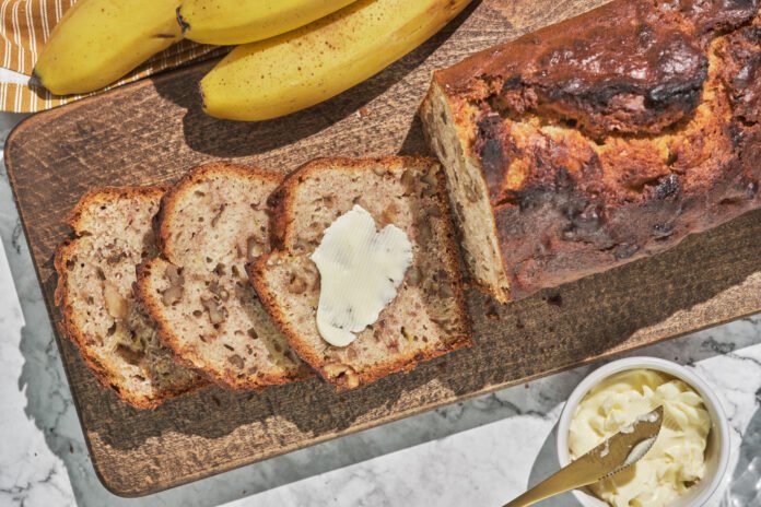 Fatias de bolo de banana e aveia com melado, servido em uma tábua de madeira ao lado de bananas frescas e manteiga.