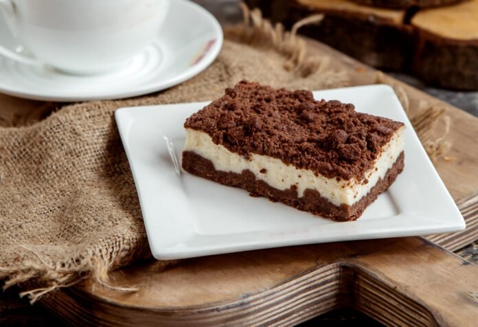 Fatia de bolo de cacau com camada de iogurte sobre um prato branco, acompanhada de uma xícara de café ao fundo.