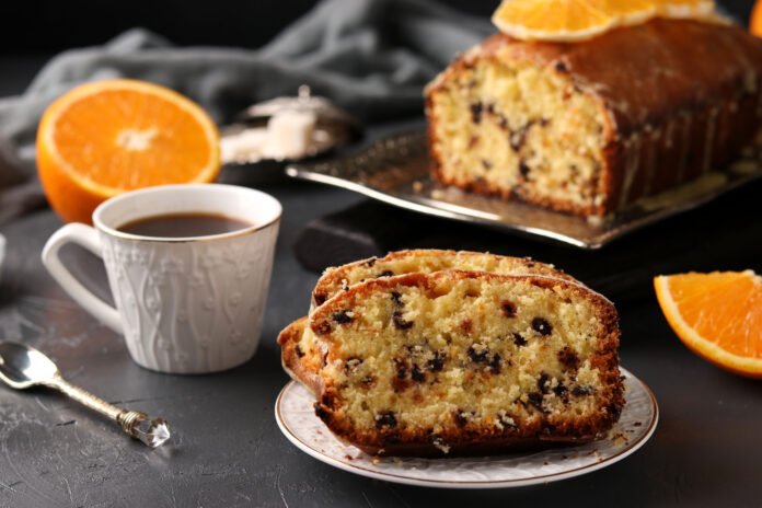 Fatia de bolo de laranja com gotas de chocolate sem açúcar servida em um prato, acompanhada de uma xícara de café e laranjas frescas ao fundo.