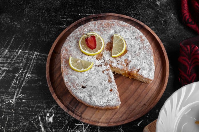 Bolo de limão fit coberto com açúcar de confeiteiro e fatias de limão decorativas em um prato de madeira.