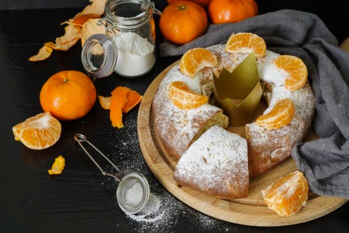 Bolo de ricota com laranja decorado com gomos de laranja, polvilhado com açúcar de confeiteiro e acompanhado por cascas de laranja, farinha e utensílios sobre uma mesa preta.