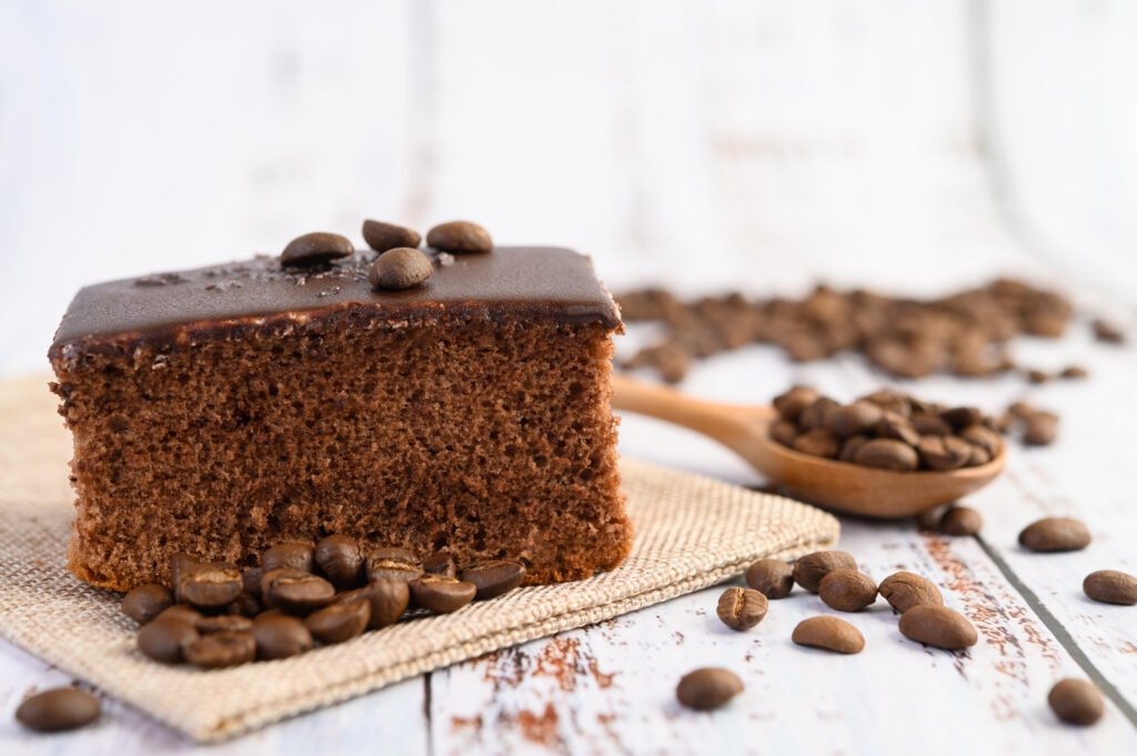 Fatia de bolo vegano de café com cobertura de chocolate, decorada com grãos de café, ao lado de uma colher de madeira com grãos espalhados.