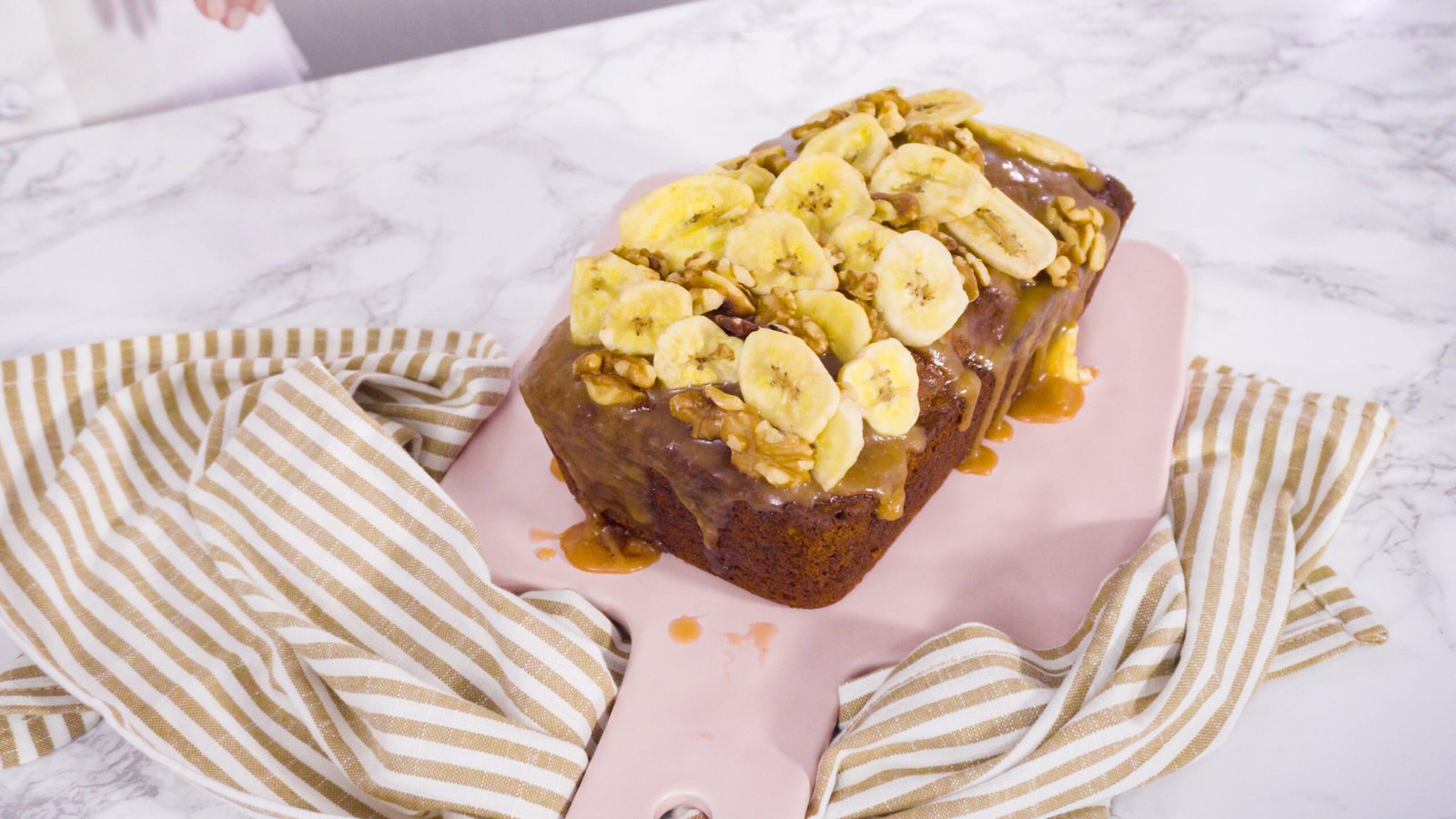 Bolo de banana e aveia com melado, decorado com fatias de banana e nozes, servido em um prato rosa com uma toalha listrada.