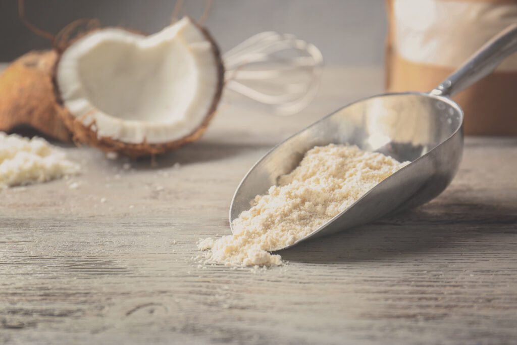 Farinha de coco em pá metálica sobre mesa de madeira, com coco aberto ao fundo.