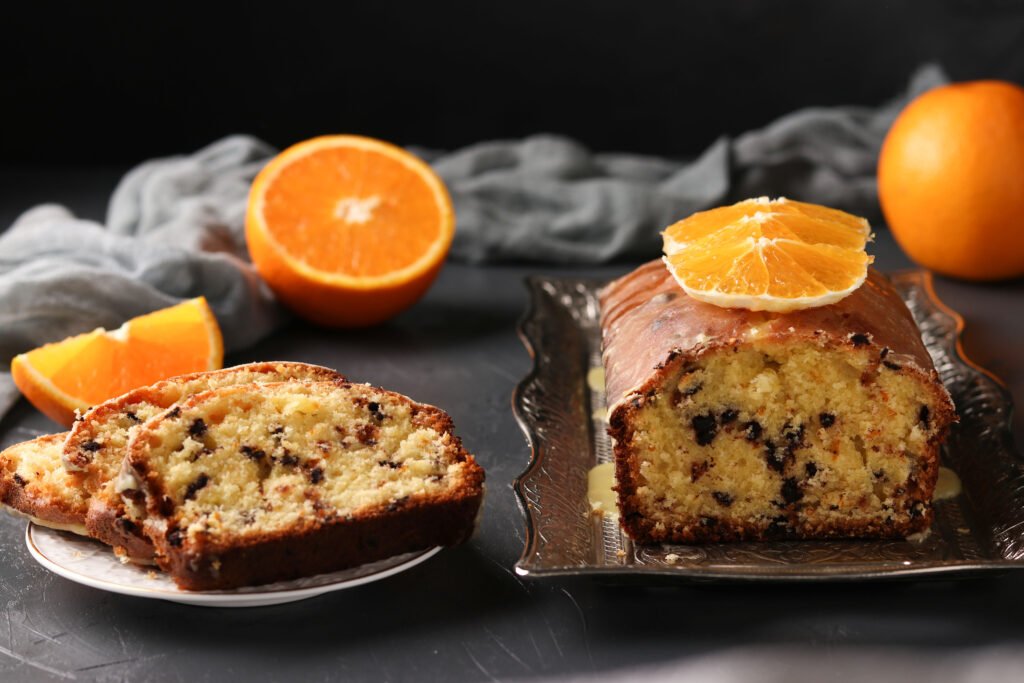 Bolo de laranja com gotas de chocolate sem açúcar, fatiado e decorado com fatias de laranja, sobre uma bandeja e um prato, acompanhado por laranjas frescas ao fundo.