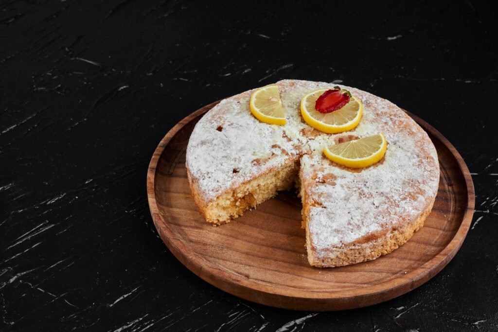 Bolo de limão fit coberto com açúcar de confeiteiro, decorado com fatias de limão e morango em um prato de madeira.