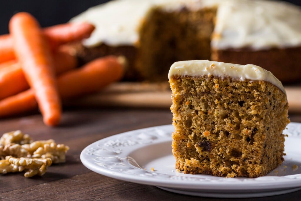 Fatia de bolo de cenoura vegano com cobertura de caramelo, decorada com nozes, ao lado de cenouras frescas em um fundo rústico.