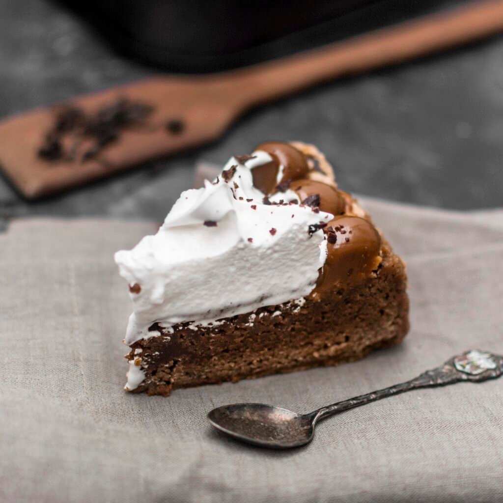 Fatia de bolo de cacau com cobertura de chantilly e doce de leite, servida sobre um guardanapo cinza, com uma colher ao lado.