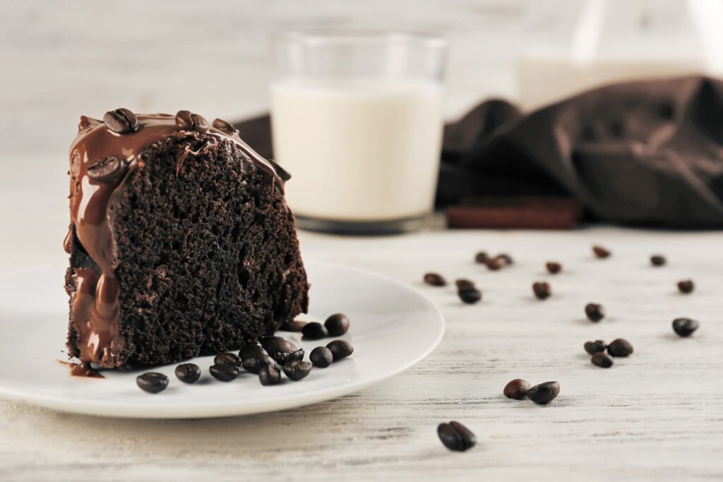 Fatia de bolo de chocolate meio amargo coberta com ganache cremosa e grãos de café decorativos, acompanhada de um copo de leite ao fundo.