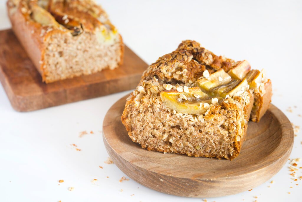 Fatia de bolo de maçã com farinha integral sem açúcar em um prato de madeira, com o bolo inteiro ao fundo em uma tábua.