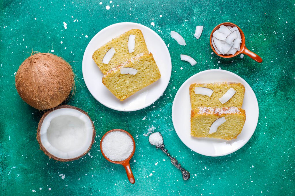 Fatias de bolo de coco com leite de amêndoas sem açúcar decoradas com coco fresco, acompanhadas de ingredientes ao fundo.