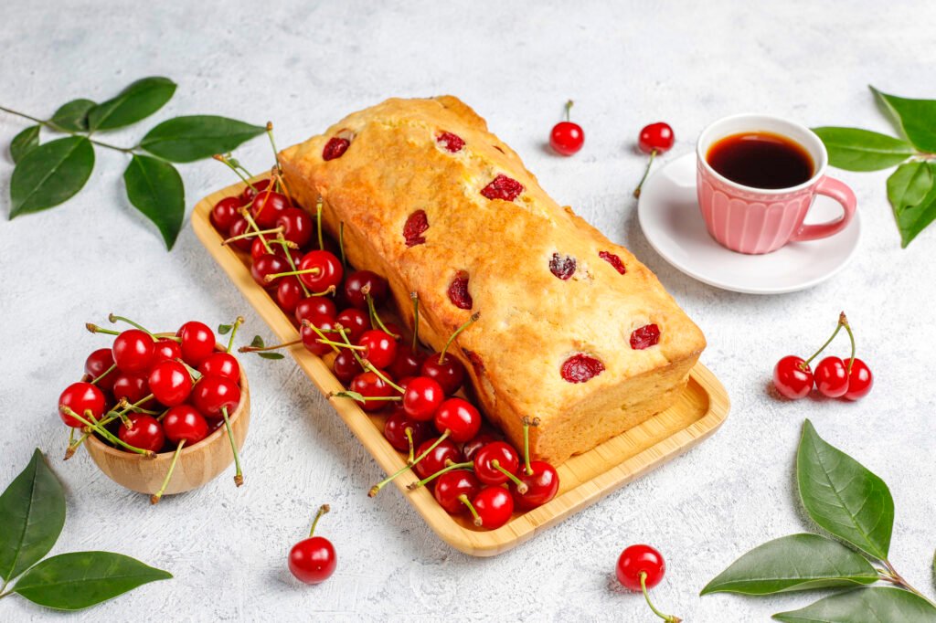 Bolo de aveia com frutas vermelhas vegano decorado com cerejas frescas, servido em uma bandeja de madeira ao lado de uma xícara de café.