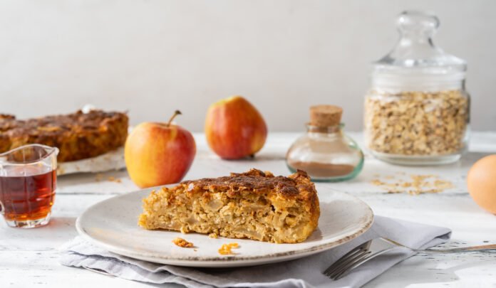 Fatia de bolo de aveia com melado em um prato branco, acompanhada de maçãs e ingredientes em segundo plano.