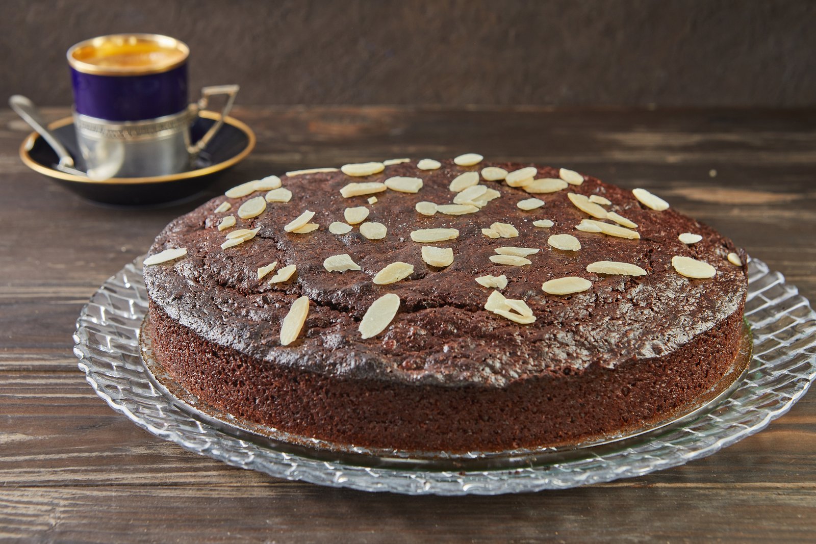 Bolo de cacau com amêndoas, decorado com fatias de amêndoas laminadas, servido em um prato de vidro com uma xícara de café ao fundo.