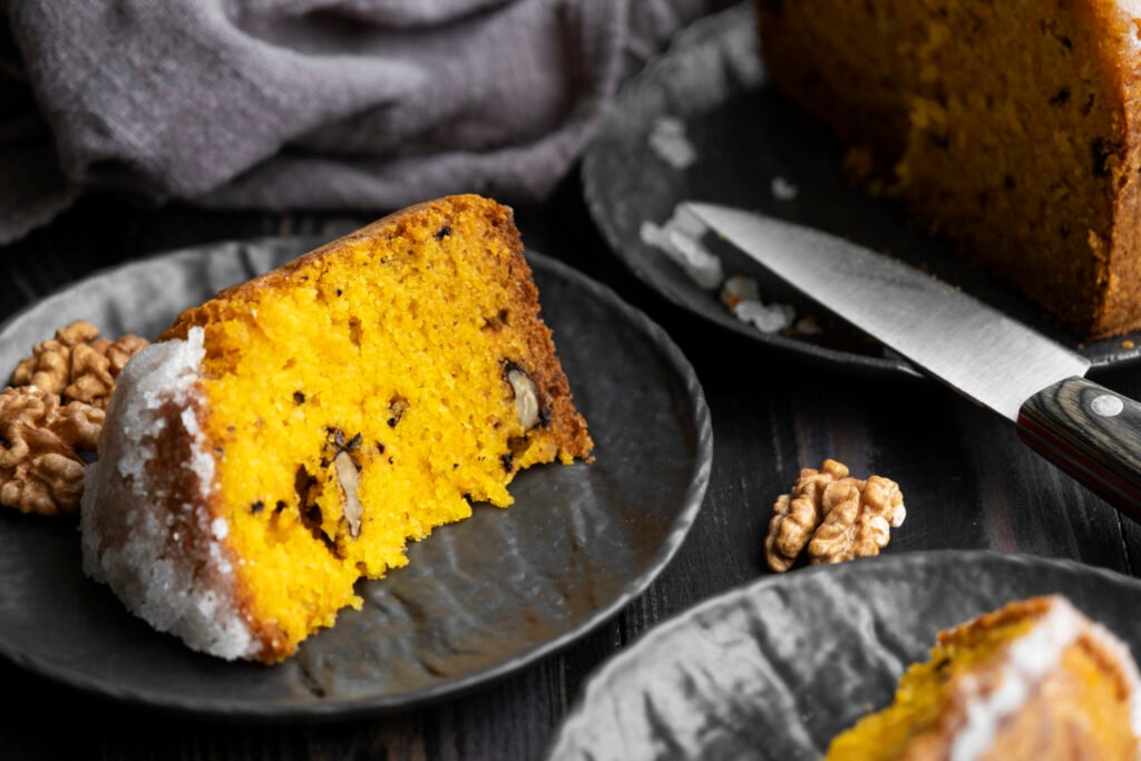 Fatia de bolo de cenoura com casca, acompanhada de nozes, em um prato escuro.