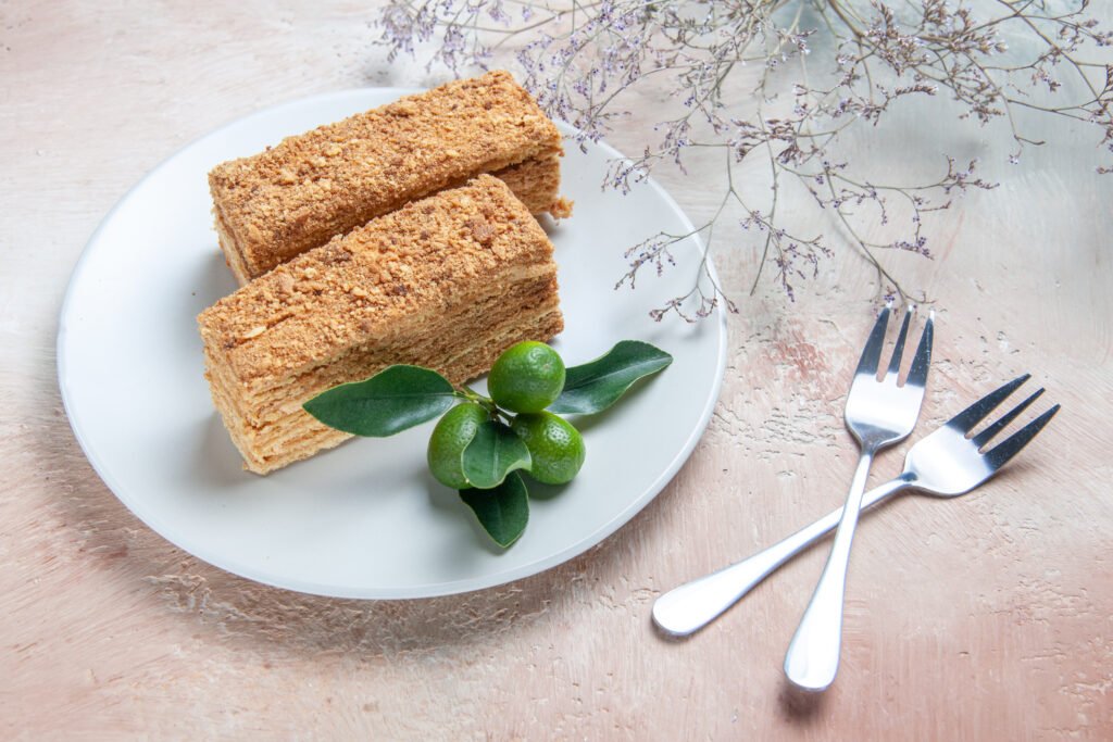 Duas fatias de bolo de fubá com coco e erva-doce servidas em um prato branco, decoradas com folhas verdes e pequenos limões, acompanhadas por dois garfos de sobremesa.