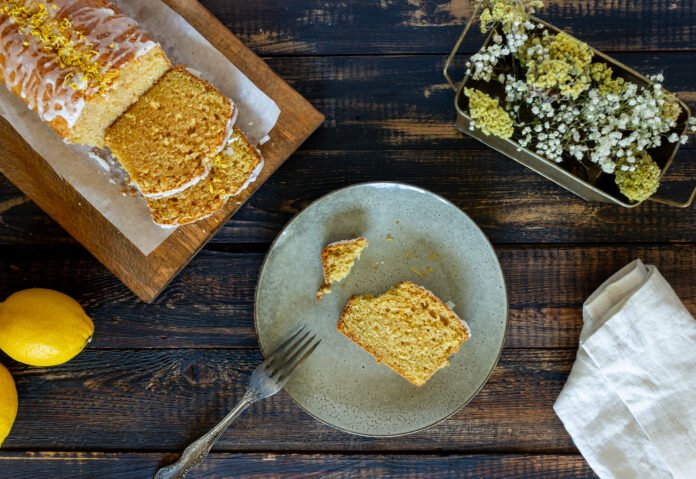 Fatia de bolo de fubá com coco e erva-doce servida em um prato de cerâmica ao lado de limões e flores decorativas sobre uma mesa rústica.