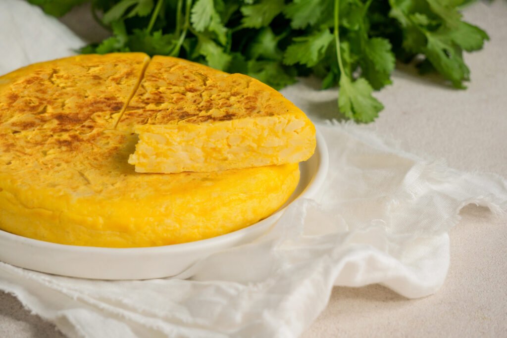 Bolo de fubá com queijo Minas servido em um prato branco, com uma fatia destacada mostrando sua textura macia, acompanhado de folhas de coentro ao fundo.