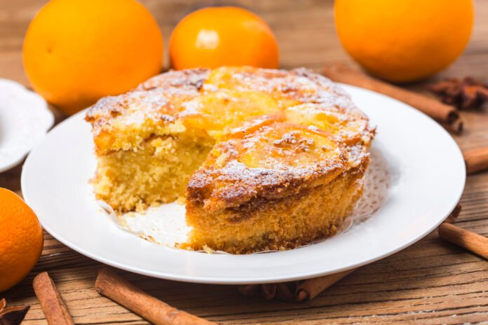 Fatia de um bolo de laranja com gengibre, decorado com fatias de laranja e açúcar de confeiteiro, em um prato branco.