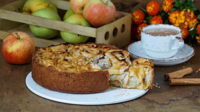 Bolo de maçã com canela e mel em um prato branco, acompanhado de xícara de chá, maçãs frescas e canela em pau.