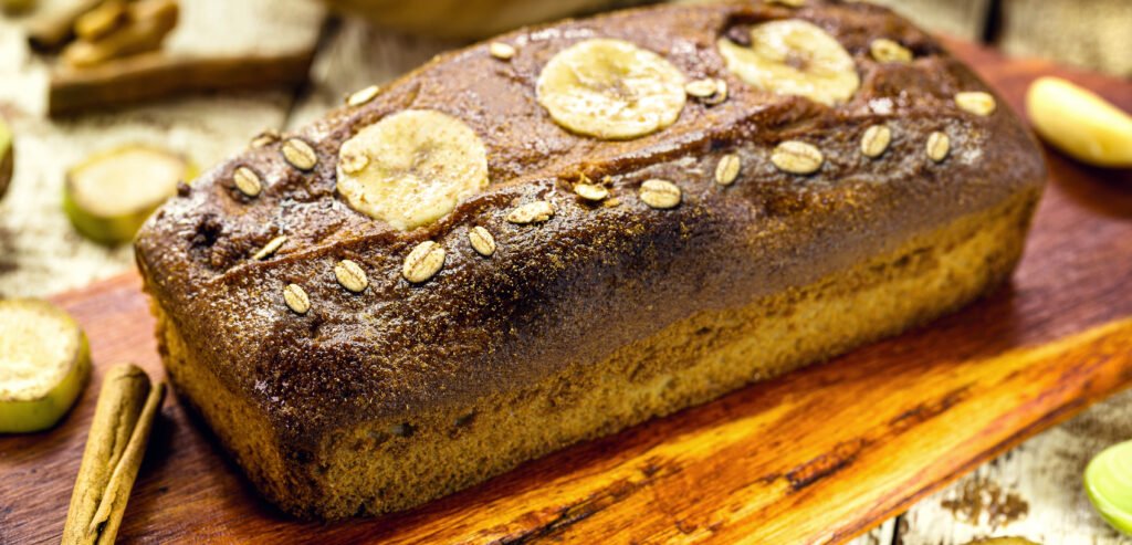 Bolo de banana com aveia decorado com fatias de banana e aveia sobre uma tábua de madeira.