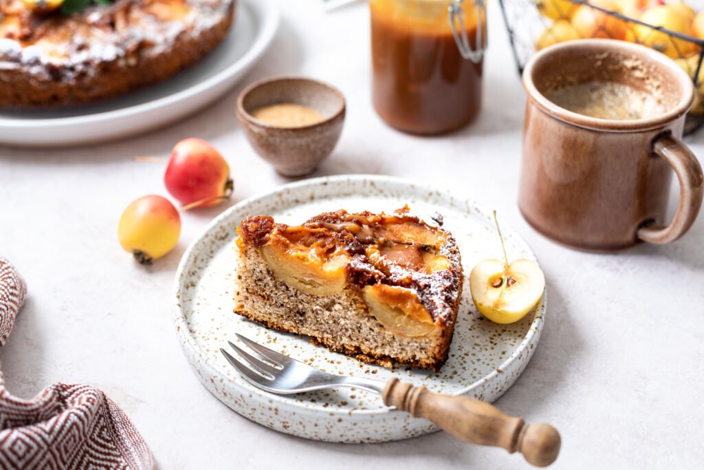 Fatia de bolo de maçã com canela e mel em prato de cerâmica, acompanhada de xícara de café e maçãs decorativas.